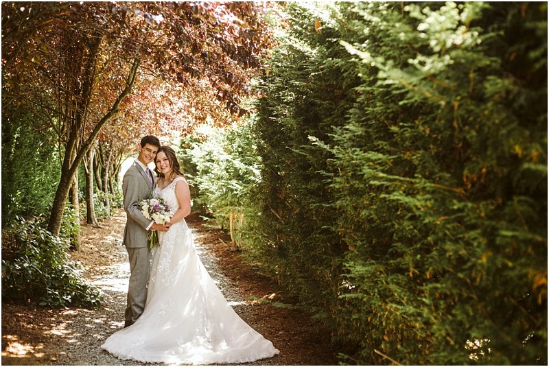 A bride in a white gown and a groom in a light gray suit smile and embrace on a tree lined path in snohomish surrounded by lush greenery and sunlight filtering through the leaves Snohomish wedding photography