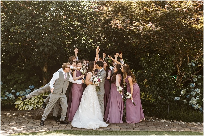 A bride in a white dress surrounded by bridesmaids in mauve and a groomsman in a gray suit all smiling and raising their hands joyfully in a lush snohomish garden filled with greenery and flowers Snohomish wedding photography