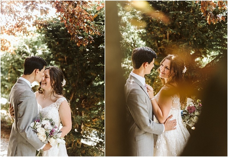 A bride and groom stand outdoors in snohomish smiling and gazing at each other lovingly The bride holds a bouquet of flowers and wears a white lace dress the groom is in a light gray suit Sunlight filters through the trees behind them Snohomish wedding photography