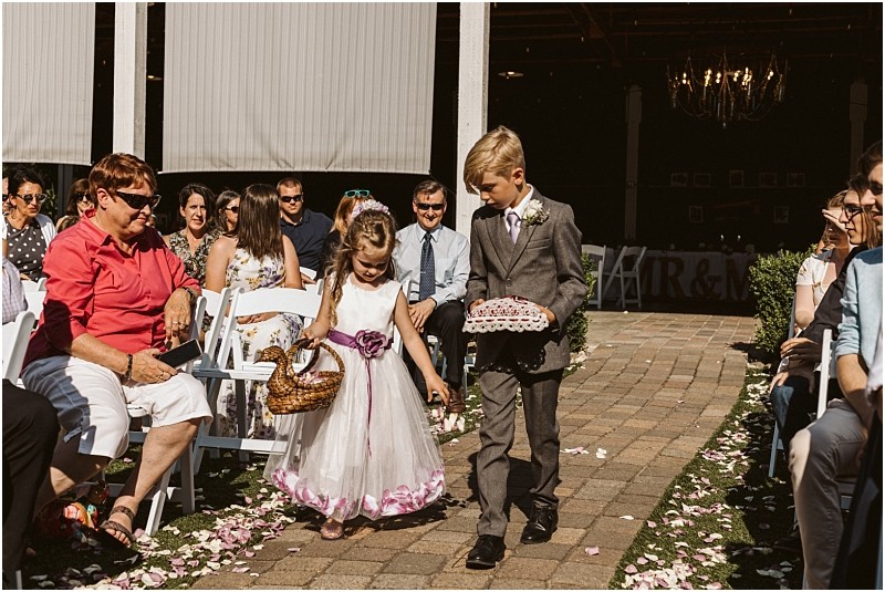 A young flower girl and ring bearer walk down a sunlit snohomish outdoor aisle lined with petals surrounded by seated guests The girl carries a basket and wears a white dress the boy wears a suit and holds a pillow Snohomish wedding photography
