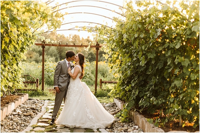 A bride and groom stand in a lush Snohomish garden walkway, facing each other and smiling lovingly. The bride wears a white gown, the groom a gray suit. Sunlight filters through greenery and a wooden arbor overhead. Snohomish Wedding Photography
