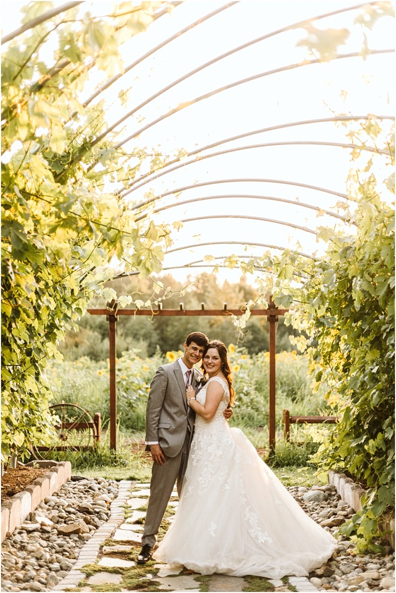 A bride and groom stand smiling and embracing under a sunlit vine covered trellis in a snohomish outdoor garden The bride wears a white gown and the groom is in a light gray suit surrounded by lush greenery Snohomish wedding photography
