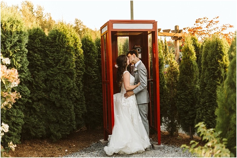 A bride and groom share a kiss inside a red phone booth surrounded by tall green bushes and trees on a sunny day in snohomish Snohomish wedding photography