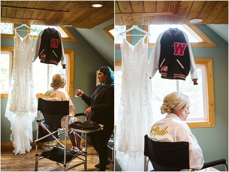 A bride with blonde hair sits in a chair wearing a white robe with bride on the back having her makeup done In a bright wood paneled snohomish room a lace wedding dress and varsity jacket hang by the window Snohomish wedding photography