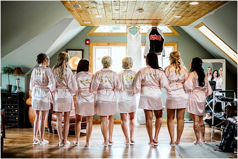 Eight women in matching pale pink robes stand with their backs to the camera in a cozy snohomish room looking at a wedding dress and a black jacket hanging from the ceiling as sunlight streams through skylights above Snohomish wedding photography
