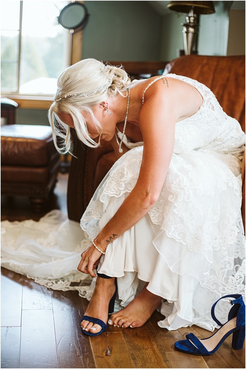A bride with blonde hair in an elegant lace wedding dress sits on a leather chair in snohomish leaning forward to fasten the strap on a blue high heeled sandal with natural light streaming in from a nearby window Snohomish wedding photography