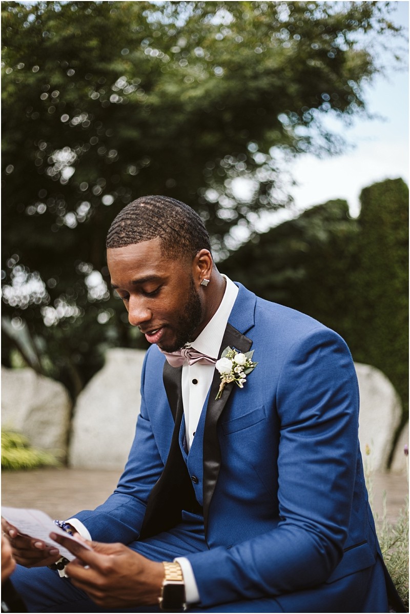 A man in a blue suit with a boutonniere sits outdoors in snohomish reading a letter and smiling softly Trees and large rocks are visible in the background Snohomish wedding photography