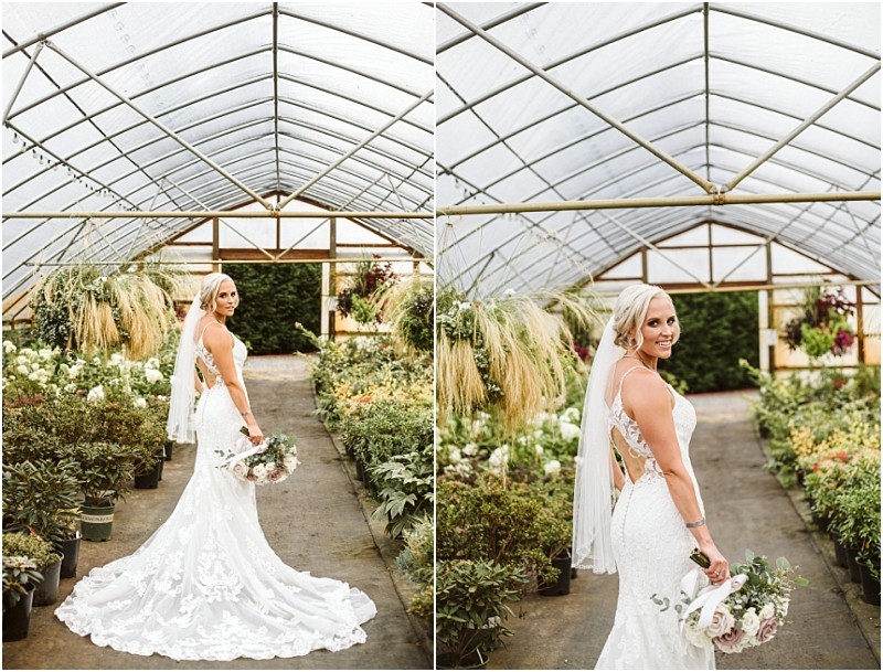 A bride in a white lace gown and veil holds a bouquet and poses in a snohomish greenhouse surrounded by lush green plants and flowers She smiles over her shoulder standing on a concrete path Snohomish wedding photography