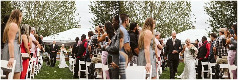 A bride in a white dress walks down an outdoor aisle with an older man in snohomish surrounded by standing guests under a cloudy sky with trees and a white tent in the background Snohomish wedding photography