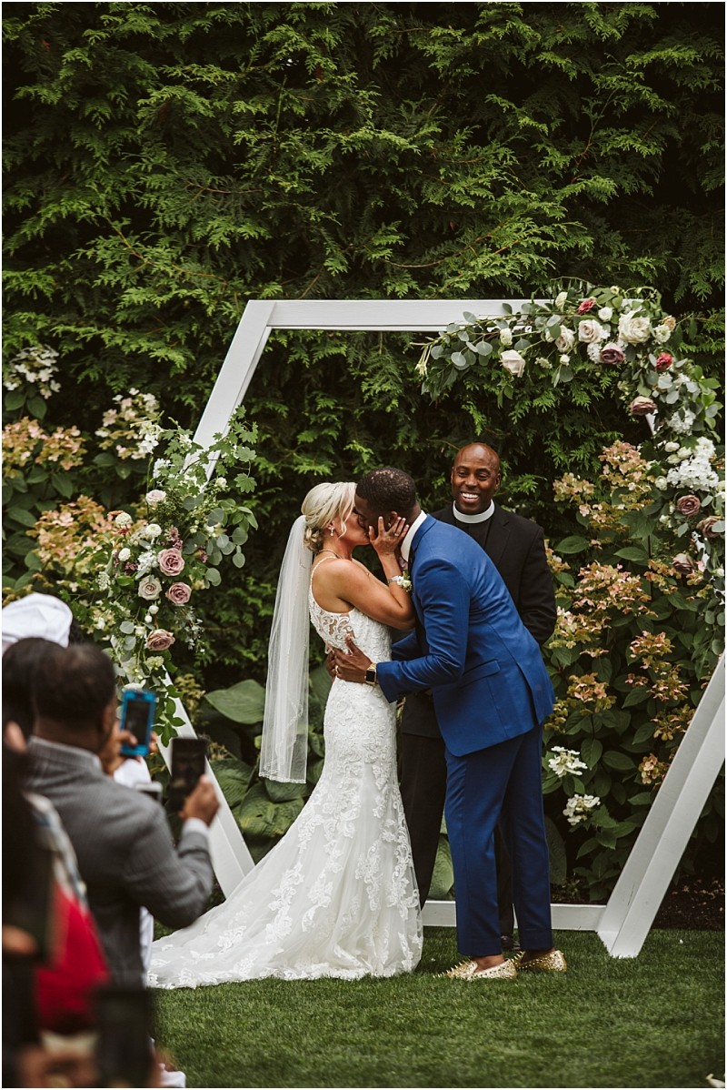 A bride and groom kiss at their outdoor snohomish wedding ceremony under a geometric floral arch with a smiling officiant behind them and guests taking photos in the foreground Snohomish wedding photography