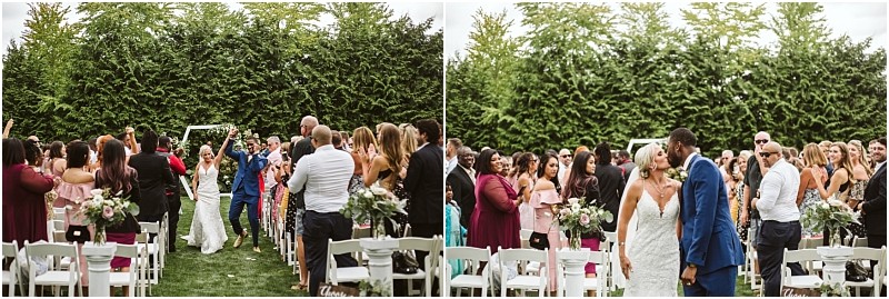A bride in a white gown and a groom in a blue suit walk down an outdoor snohomish aisle surrounded by standing guests some taking photos and clapping with greenery and trees in the background Snohomish wedding photography