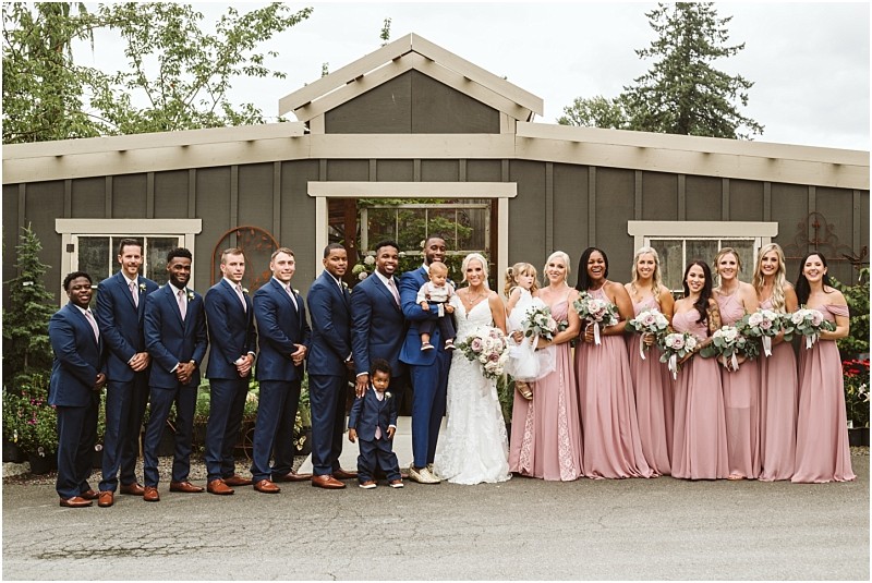 A wedding party poses in front of a rustic snohomish building The group includes the bride groom bridesmaids in blush pink dresses groomsmen in navy suits and two young boys all smiling and holding bouquets or hands Snohomish wedding photography