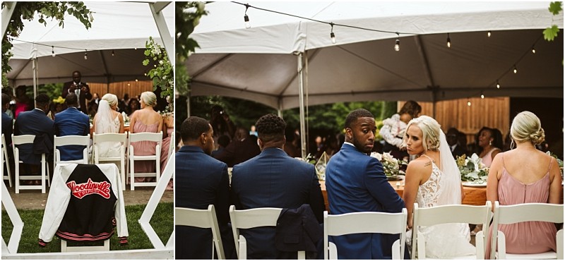 Wedding guests and a bride and groom sit under a white tent at an outdoor reception in snohomish A varsity jacket with woodinville on the back hangs on a chair in the foreground while the bride looks back toward the camera Snohomish wedding photography