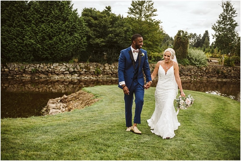 A bride in a white dress and veil holds a bouquet while walking hand in hand with a groom in a blue suit on green grass by a pond in snohomish both smiling and surrounded by trees and lush greenery Snohomish wedding photography