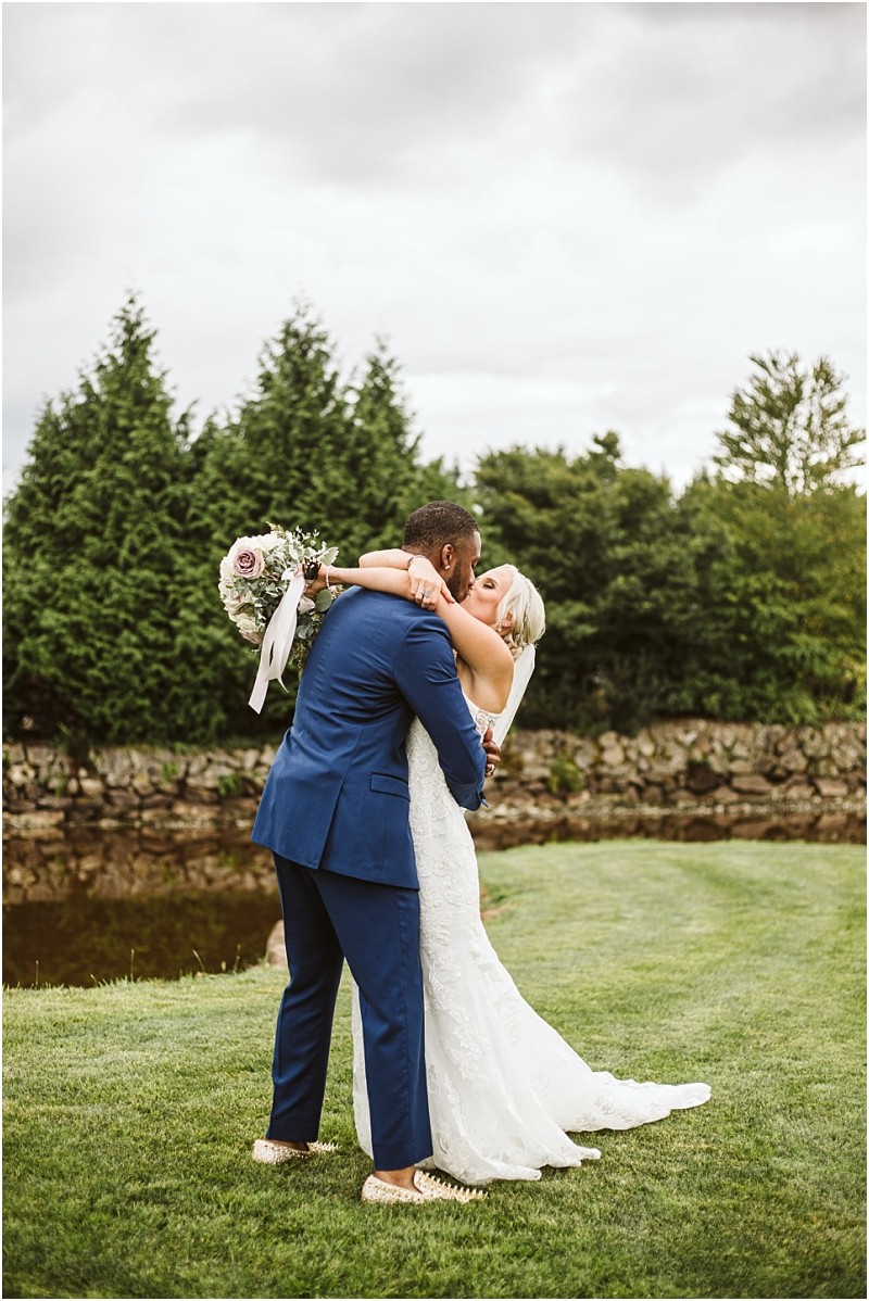 A bride in a white gown embraces a groom in a blue suit outdoors on green grass in snohomish with trees and a stone wall behind them The bride holds a bouquet of pink and white flowers Snohomish wedding photography