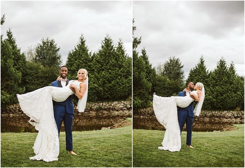 A groom in a blue suit stands on grass holding his bride in a white gown in his arms They smile and pose for photos outdoors in charming snohomish with trees and a pond in the background under a cloudy sky Snohomish wedding photography