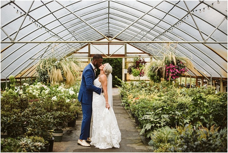 A bride and groom share a kiss inside a Snohomish greenhouse filled with lush green plants and hanging flowers. The groom wears a blue suit and the bride wears a white lace gown. Snohomish Wedding Photography
