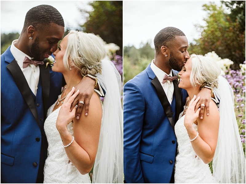 A bride and groom embrace outdoors in snohomish The groom in a blue suit gently touches foreheads and kisses the blonde bride who wears a white dress and veil Both smile surrounded by greenery and purple flowers Snohomish wedding photography