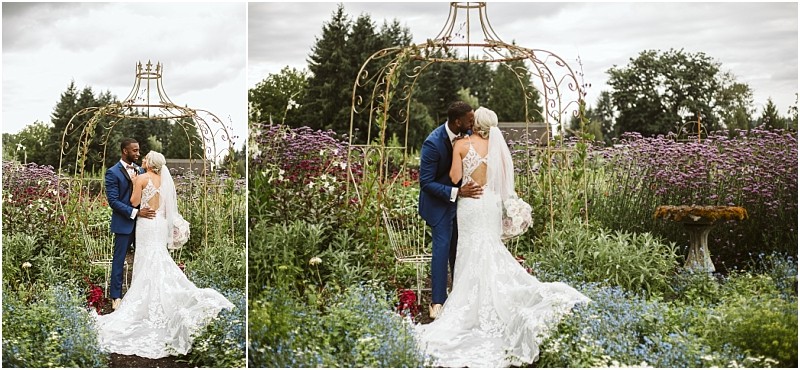 A bride in a white gown and veil embraces a groom in a blue suit in a lush snohomish garden standing before a gold arbor with blooming purple and blue flowers surrounding them Snohomish wedding photography