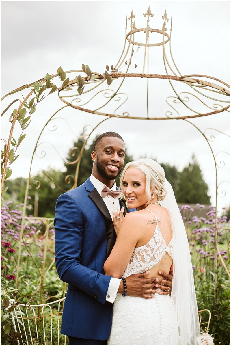 A bride in a white lace gown and a groom in a blue tuxedo smile and embrace under an ornate metal arch in a snohomish garden filled with purple flowers Snohomish wedding photography