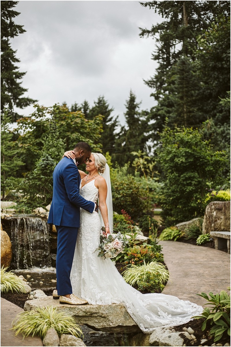 A bride and groom embrace on a garden path beside a small waterfall in snohomish The bride wears a white lace gown and veil holding a bouquet while the groom wears a blue suit Lush greenery and trees surround them under a cloudy sky Snohomish wedding photography
