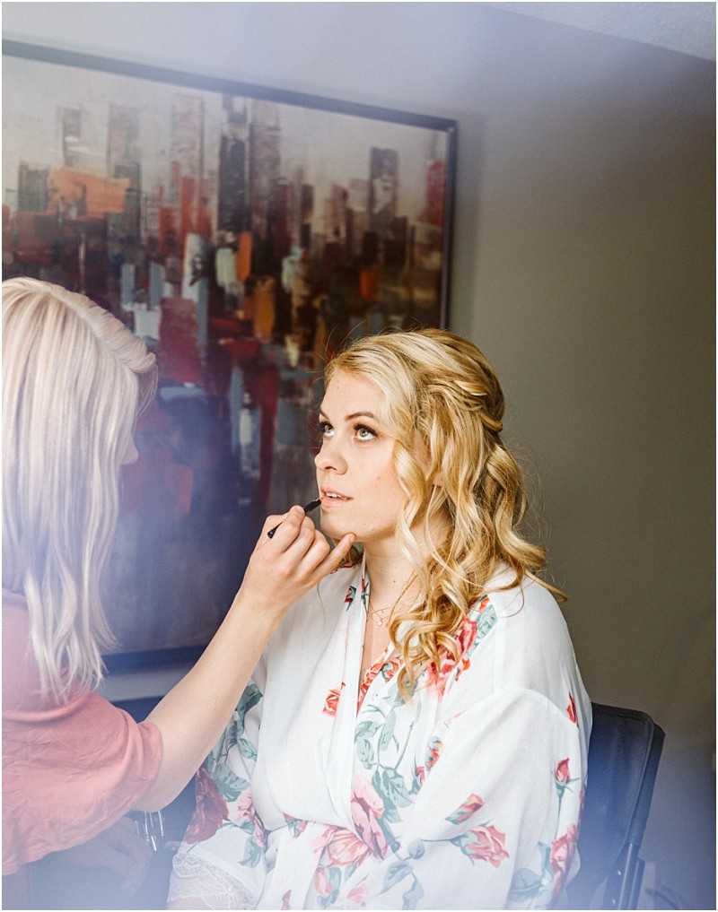 A woman with blonde hair in soft curls wears a floral robe while having her makeup done by another woman Indoors a colorful cityscape painting hints at downtown snohomish in the background Snohomish wedding photography