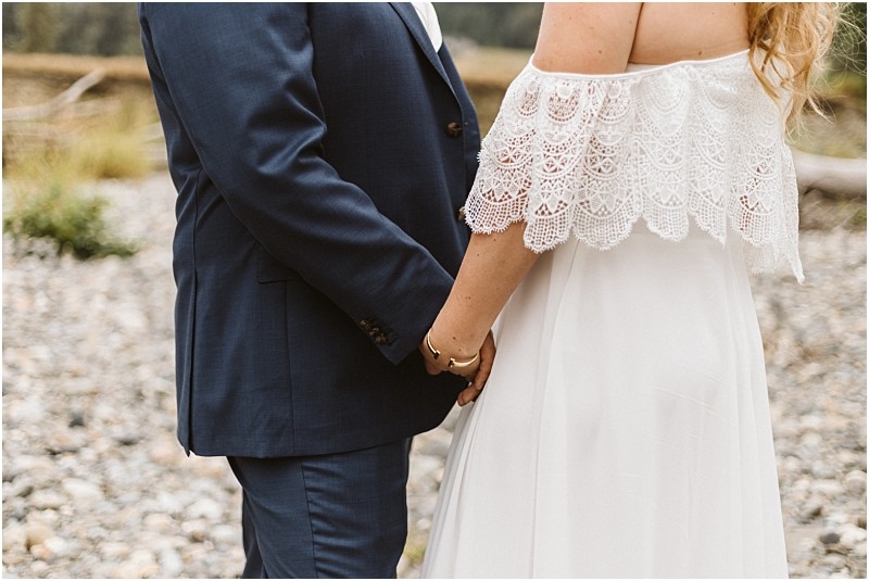 A couple holds hands outdoors in snohomish the person on the left wears a navy blue suit while the one on the right dons an off the shoulder white lace dress Only their torsos and joined hands are visible Snohomish wedding photography
