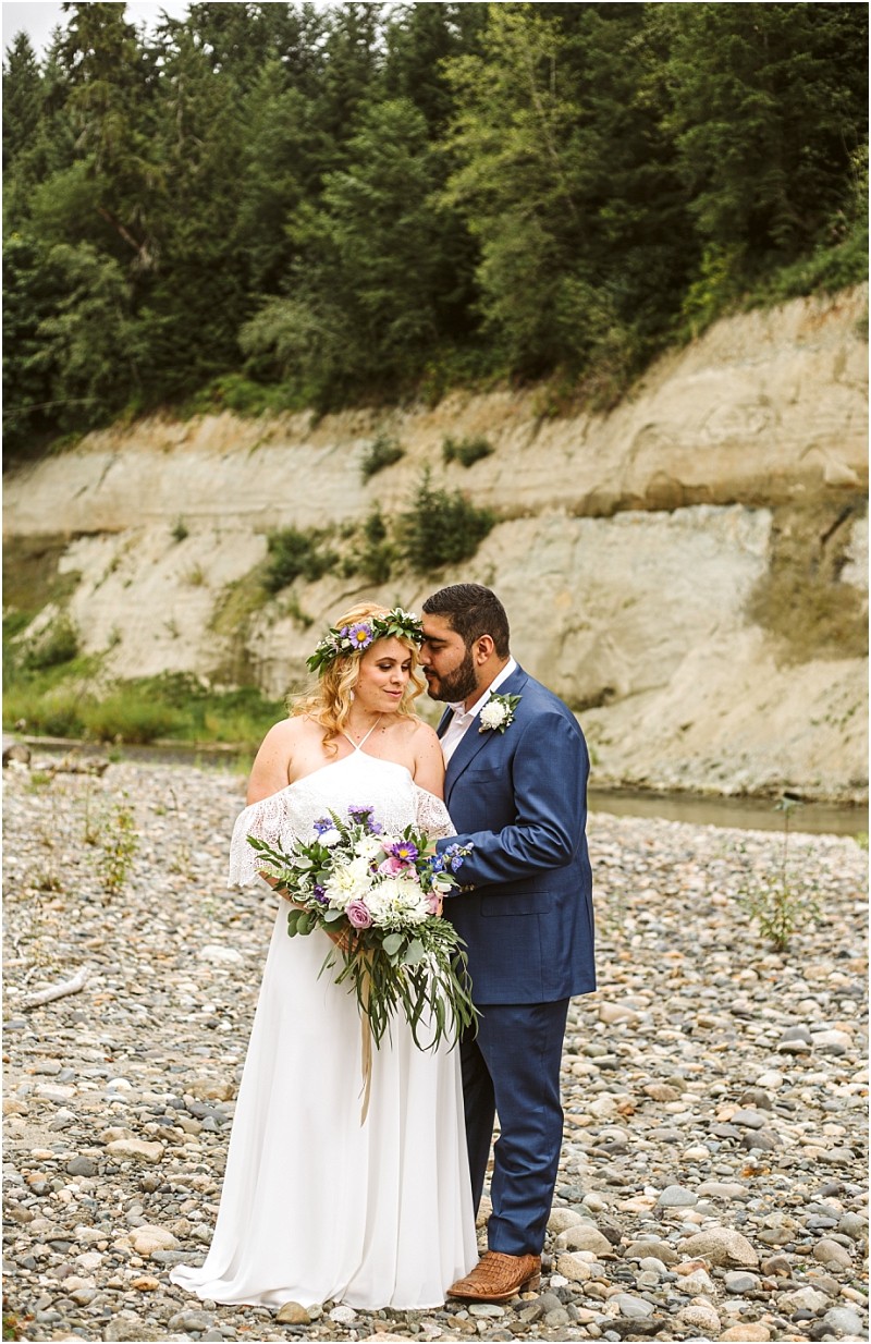 A bride in a white dress and flower crown holds a bouquet standing close to a groom in a blue suit on a rocky snohomish riverbank with trees and a cliffside in the background Snohomish wedding photography