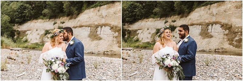 A bride with a flower crown and a groom in a blue suit stand close together smiling and laughing holding a large bouquet on a rocky snohomish riverbank with trees and a cliff in the background Snohomish wedding photography