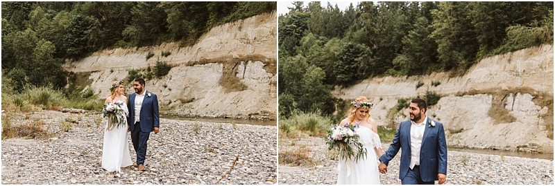 A bride in a white dress and floral crown holds a bouquet while walking hand in hand with a groom in a blue suit along a rocky snohomish riverbed with a forest and cliffside in the background Snohomish wedding photography
