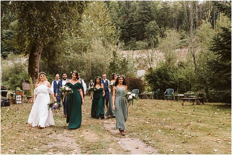 A bride in a white dress and bridesmaids in green dresses walk outdoors on a grassy path surrounded by trees with groomsmen following at a rustic snohomish forested wedding venue Snohomish wedding photography