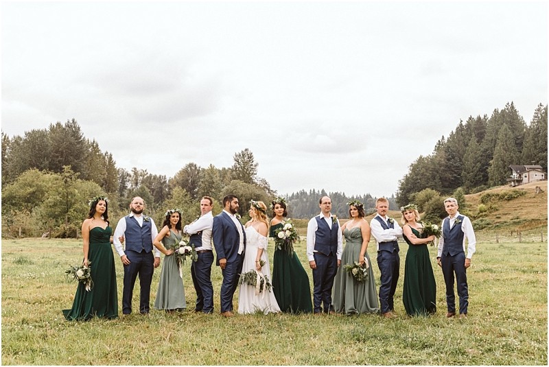 A wedding party poses outdoors on a grassy field in snohomish with trees and hills in the background The bridesmaids wear various shades of green dresses and the groomsmen wear blue vests or jackets with white shirts and dark pants Snohomish wedding photography
