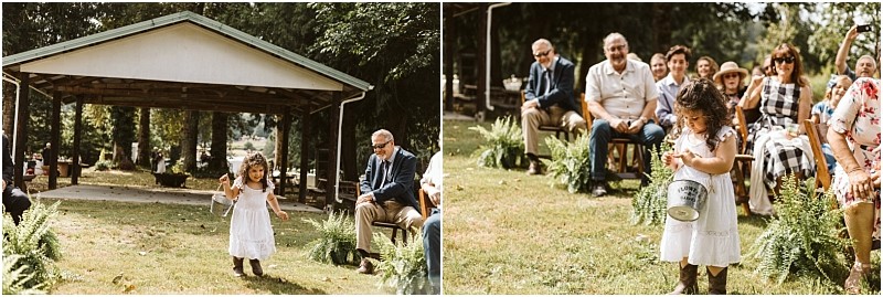 A young girl in a white dress and boots walks outside at a snohomish wedding holding a pail Guests seated on benches watch her and smile surrounded by lush greenery Snohomish wedding photography