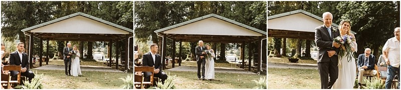 A bride walks down an outdoor aisle with two men in snohomish approaching a small pavilion Guests sit on wooden chairs on either side surrounded by trees and greenery on a sunny day Snohomish wedding photography