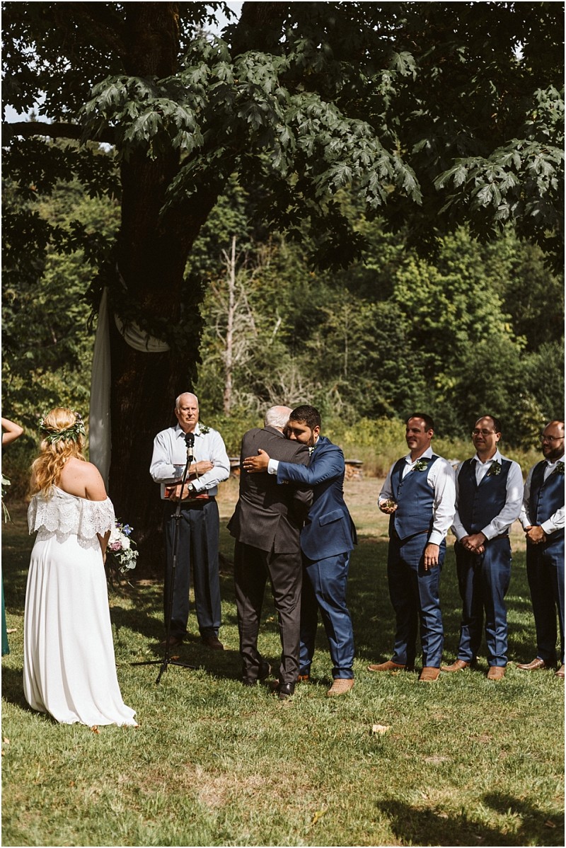 A bride in a white dress stands at a snohomish outdoor wedding ceremony as two men embrace under a large tree groomsmen in blue suits watch and an officiant stands nearby Snohomish wedding photography
