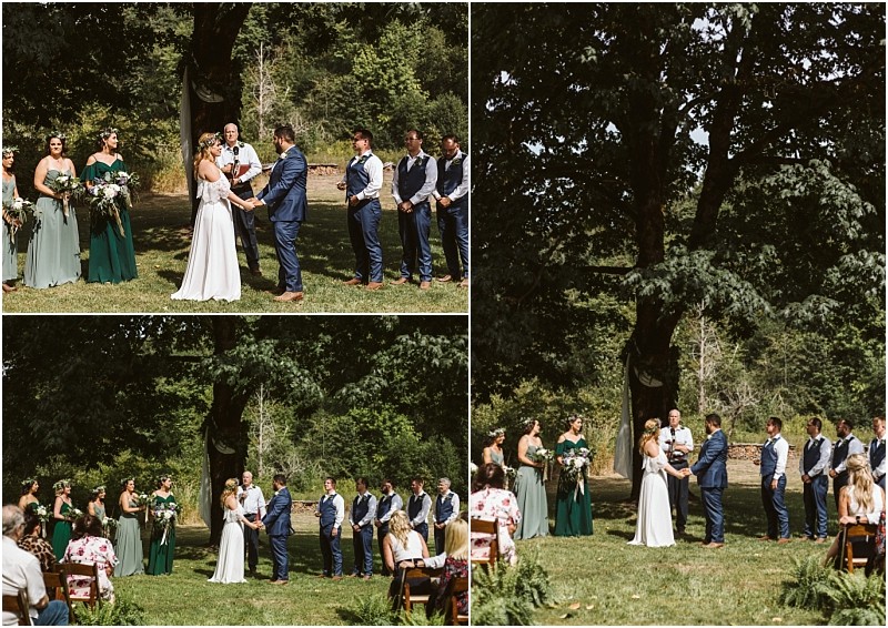 A beautiful outdoor wedding ceremony in snohomish shows a bride and groom exchanging vows under a large tree surrounded by bridesmaids in green dresses and groomsmen in blue suits with guests seated on the lawn Snohomish wedding photography