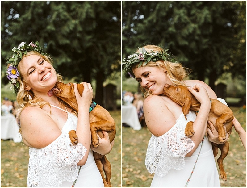 A smiling woman in a white dress and flower crown cuddles a brown puppy outdoors in snohomish The puppy playfully licks her face as she laughs surrounded by greenery in a joyful candid moment Snohomish wedding photography