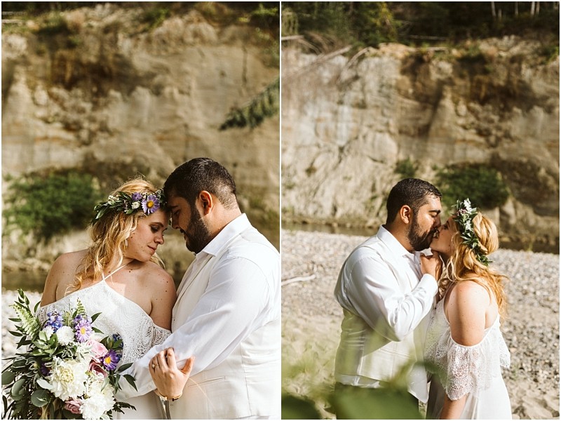 A couple in wedding attire embraces outdoors near snohomishs rocky cliffs and greenery The bride wears a flower crown and white dress holding a bouquet while the groom is in a white suit They share intimate loving moments Snohomish wedding photography