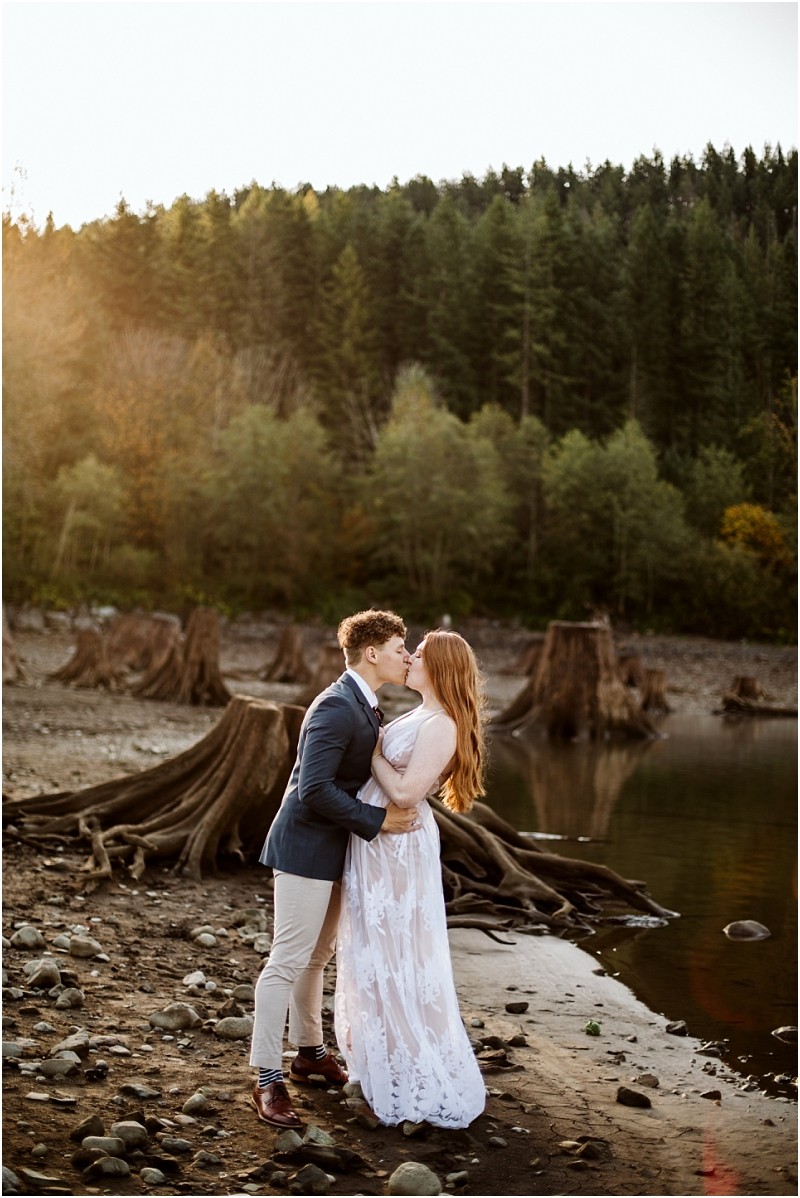 Premier snohomish & seattle wedding photographer | gsquared weddings photography a couple dressed in formal attire embraces and kisses on a rocky lakeshore near snohomish with tree stumps and a dense pine forest in the background The soft light suggests either sunrise or sunset capturing a romantic seattle wedding engagement moment | serving snohomish seattle & beyond Premier snohomish & seattle wedding photographer | gsquared weddings photography a couple dressed in formal attire embraces and kisses on a rocky lakeshore near snohomish with tree stumps and a dense pine forest in the background The soft light suggests either sunrise or sunset capturing a romantic seattle wedding engagement moment | serving snohomish seattle & beyond