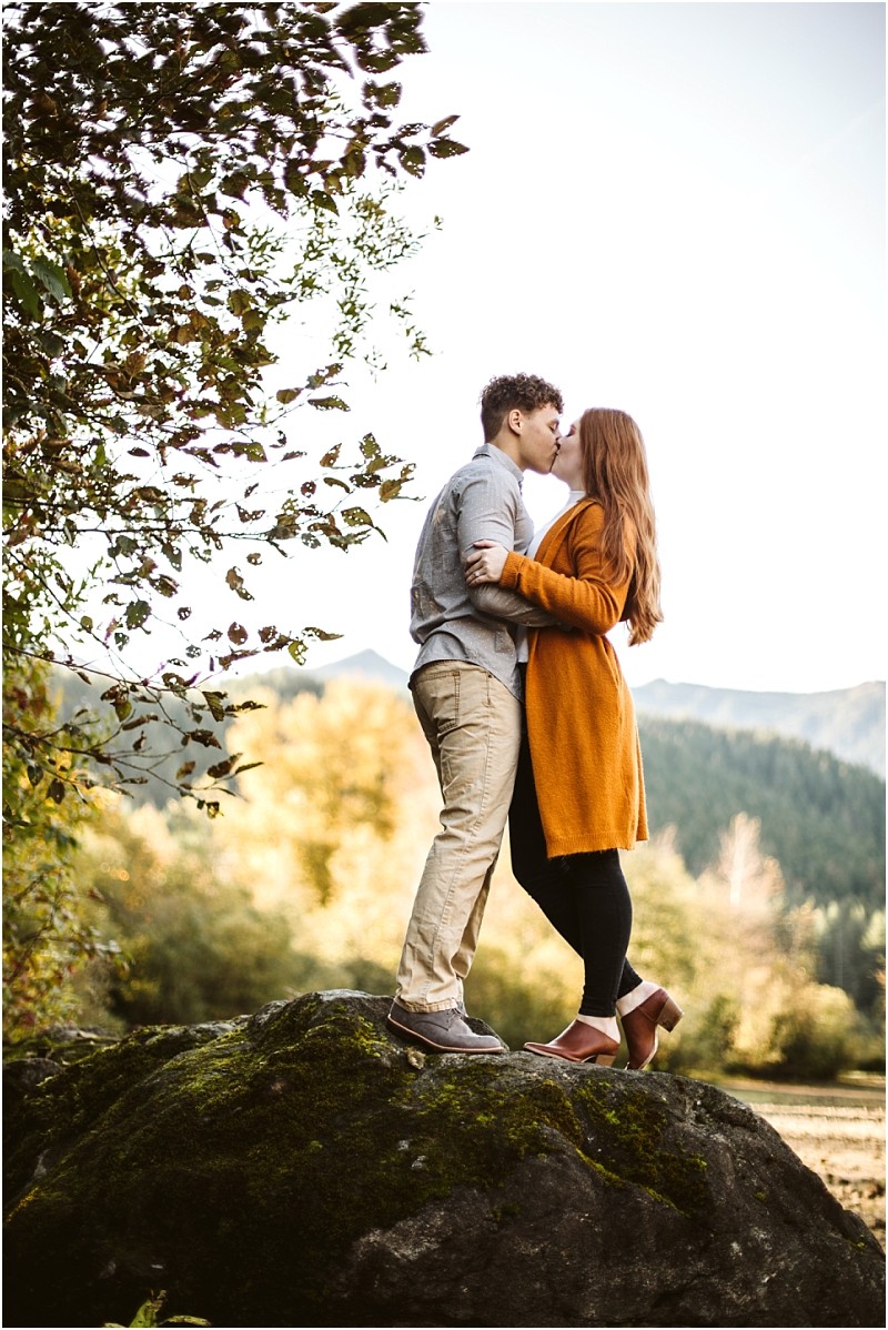 Premier snohomish & seattle wedding photographer | gsquared weddings photography a couple stands on a large mossy rock embracing and kissing outdoors during their snohomish engagement The woman wears a long orange coat and the man wears a gray shirt and khaki pants with trees and mountains in the background | serving snohomish seattle & beyond Premier snohomish & seattle wedding photographer | gsquared weddings photography a couple stands on a large mossy rock embracing and kissing outdoors during their snohomish engagement The woman wears a long orange coat and the man wears a gray shirt and khaki pants with trees and mountains in the background | serving snohomish seattle & beyond