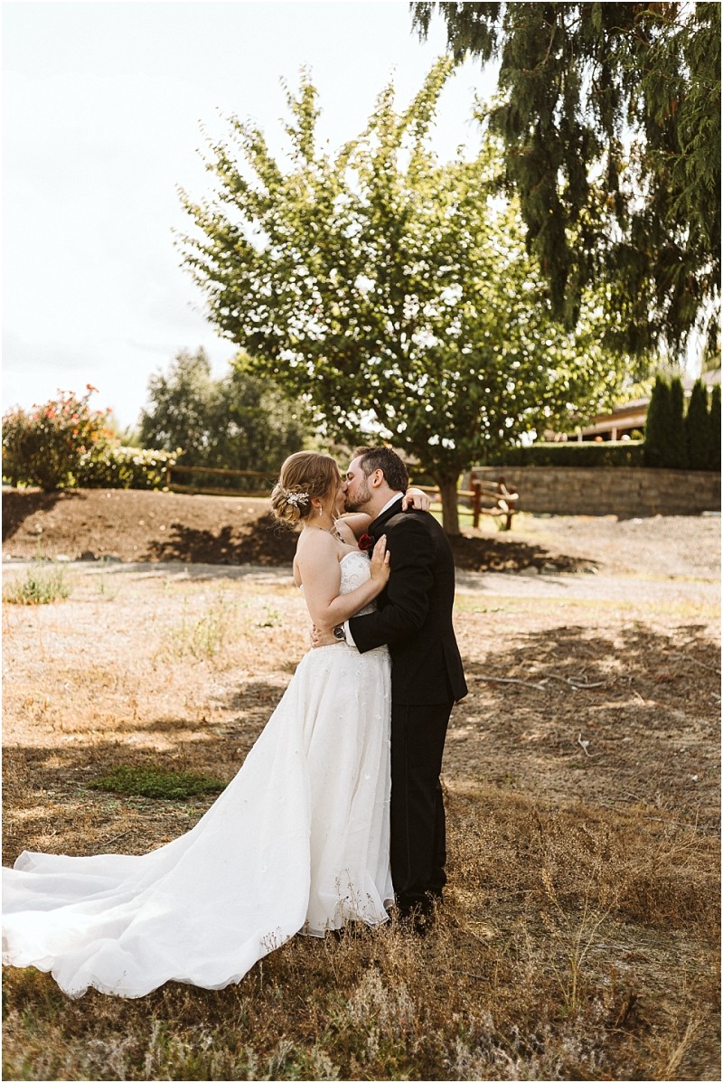 Premier snohomish & seattle wedding photographer | gsquared weddings photography a snohomish bride and groom share a kiss outdoors on a sunny day The bride wears a white strapless gown and the groom is in a black suit They stand in a grassy area with trees and a stone wall behind them at their seattle wedding | serving snohomish seattle                   & beyond