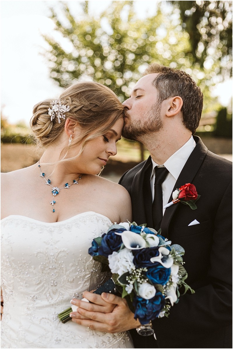 Premier snohomish & seattle wedding photographer | gsquared weddings photography a groom in a black suit gently kisses the forehead of his bride in a strapless white dress holding blue and white flowers This romantic moment is captured outdoors amid lush greenery at their snohomish seattle wedding engagement | serving snohomish seattle                   & beyond