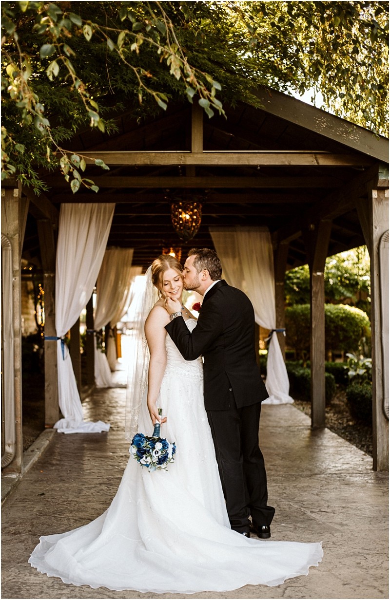 Premier snohomish & seattle wedding photographer | gsquared weddings photography a bride in a white gown holds a blue and white bouquet while a groom in a black suit kisses her cheek They stand under a wooden pergola draped with white fabric surrounded by greenery and soft sunlight at their snohomish wedding | serving snohomish seattle                   & beyond