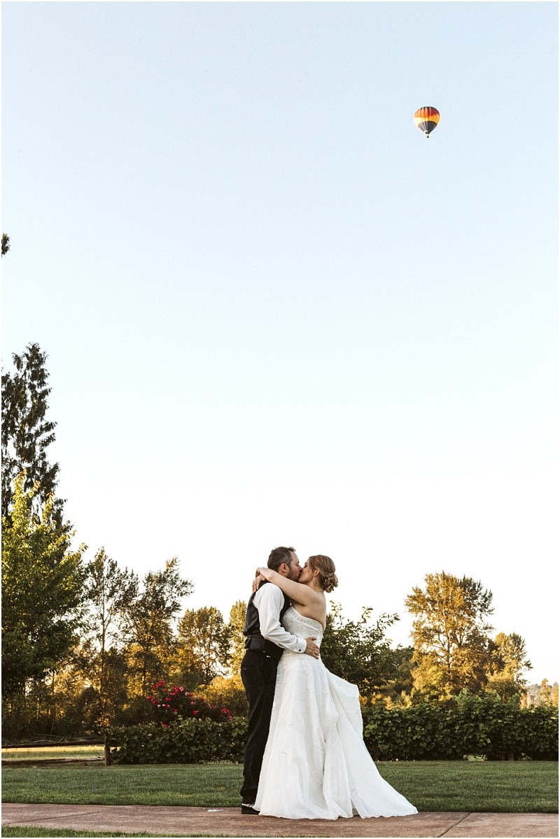 Premier snohomish & seattle wedding photographer | gsquared weddings photography a bride and groom embrace and kiss outdoors on a sunny day in snohomish surrounded by greenery with a colorful hot air balloon floating in the clear blue sky above thema dreamy seattle wedding engagement moment | serving snohomish seattle                   & beyond
