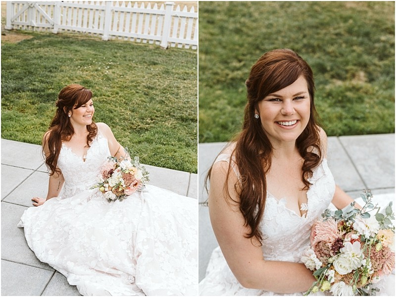 Premier snohomish & seattle wedding photographer | gsquared weddings photography a bride with long brown hair in a white lace wedding dress sits outdoors on a stone patio holding a bouquet and smiling brightly The scene captures the charm of a snohomish seattle wedding with a grassy lawn and white picket fence behind her | serving snohomish seattle                   & beyond
