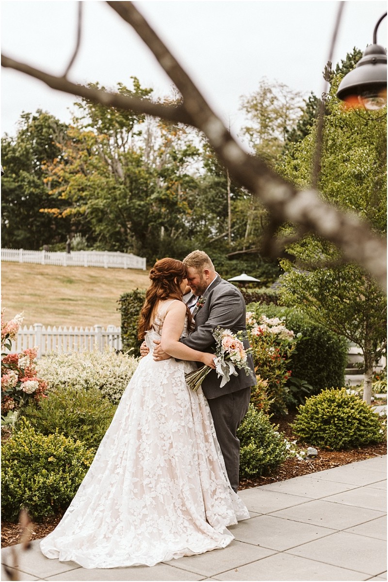 Premier snohomish & seattle wedding photographer | gsquared weddings photography a bride and groom embrace outdoors on a paved patio surrounded by lush greenery and flowers at their snohomish wedding The bride wears a white lace gown and holds a bouquet while the groom wears a gray suit Trees and a white fence are in the background | serving snohomish seattle                   & beyond