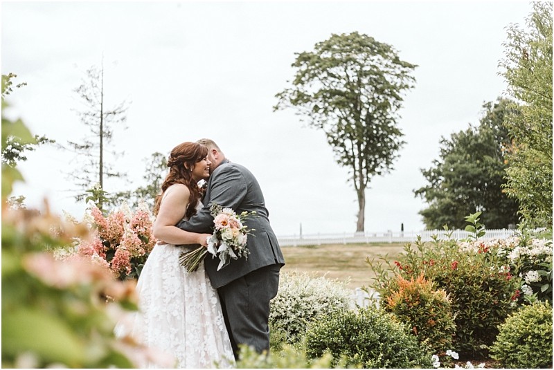 Premier snohomish & seattle wedding photographer | gsquared weddings photography a bride and groom embrace in a garden surrounded by flowers and greenery during their snohomish wedding with a large tree and open field in the background on a cloudy day | serving snohomish seattle                   & beyond