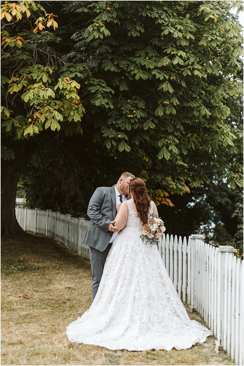Premier snohomish & seattle wedding photographer | gsquared weddings photography a bride and groom share a kiss outdoors by a white picket fence at their snohomish wedding with the bride in a lace gown holding a bouquet and the groom in a gray suit Large green trees provide shade in the background | serving snohomish seattle                   & beyond