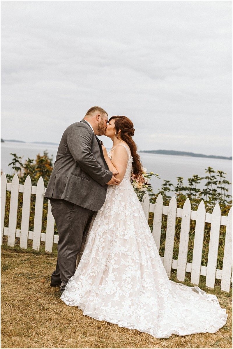 Premier snohomish & seattle wedding photographer | gsquared weddings photography a bride and groom share a kiss outdoors by a white picket fence with water and greenery in the background during their snohomish wedding The bride wears a lace gown while the groom is in a gray suit | serving snohomish seattle                   & beyond