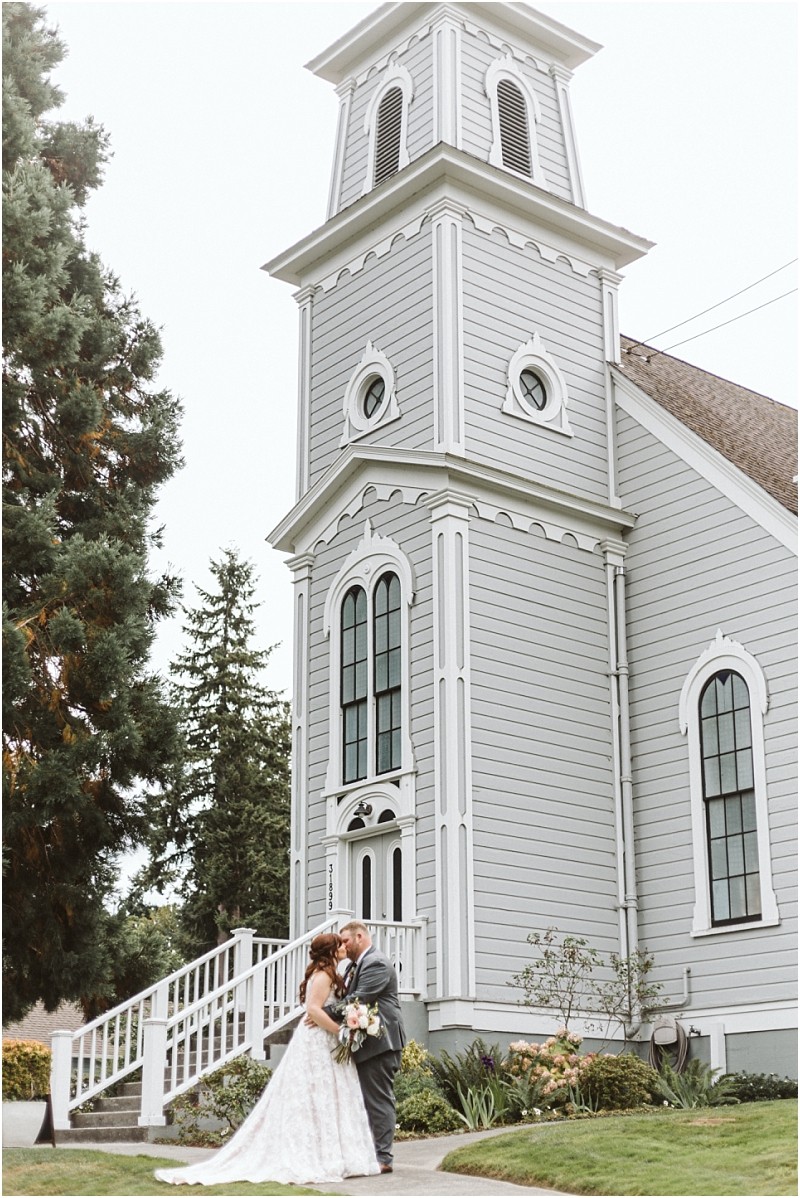 Premier snohomish & seattle wedding photographer | gsquared weddings photography a bride and groom stand together looking at each other outside a tall light gray wooden church with large arched windows and white trim in snohomish surrounded by greenery on a cloudy seattle wedding day | serving snohomish seattle                   & beyond